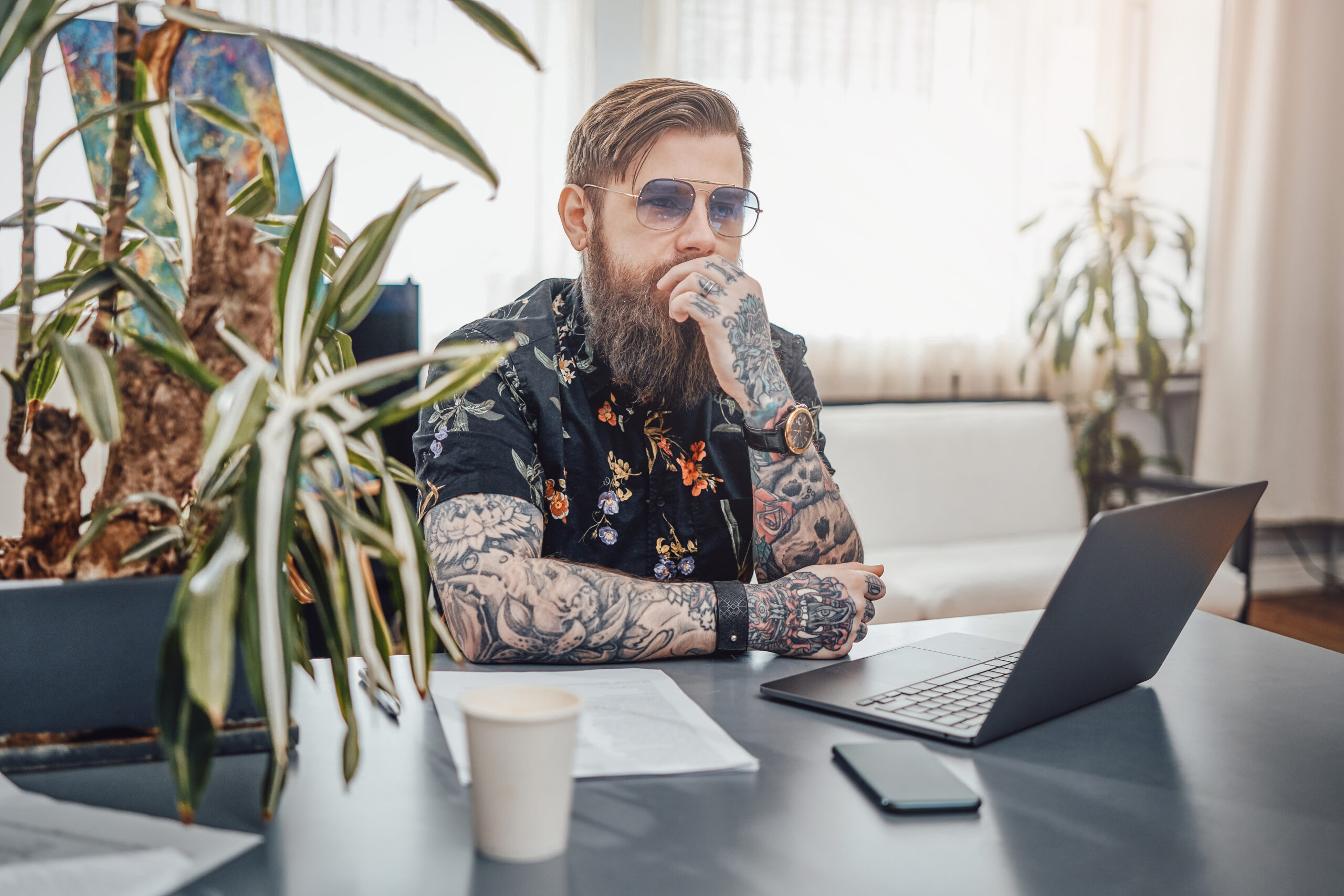 Bearded freelancer sits at table with plant and laptop in cosy room Beautiful interior of tattoo master workplace in modern studio